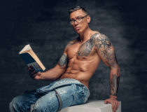 Shirtless muscular male with tattoo on a chest sits on a white wooden box and reads a book.