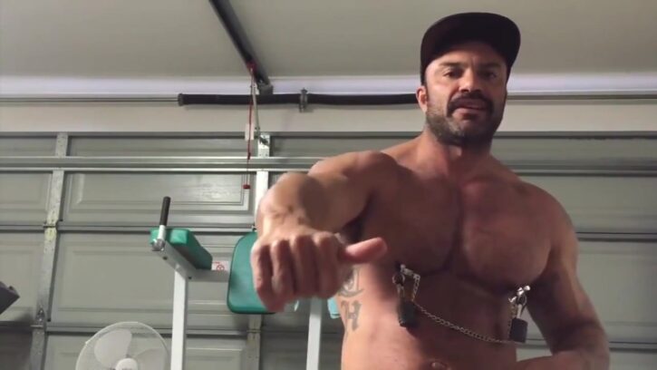 gay porn content creator and actor rogan richards standing naked in his home gym wearing black baseball cap and large metal nipple clamps