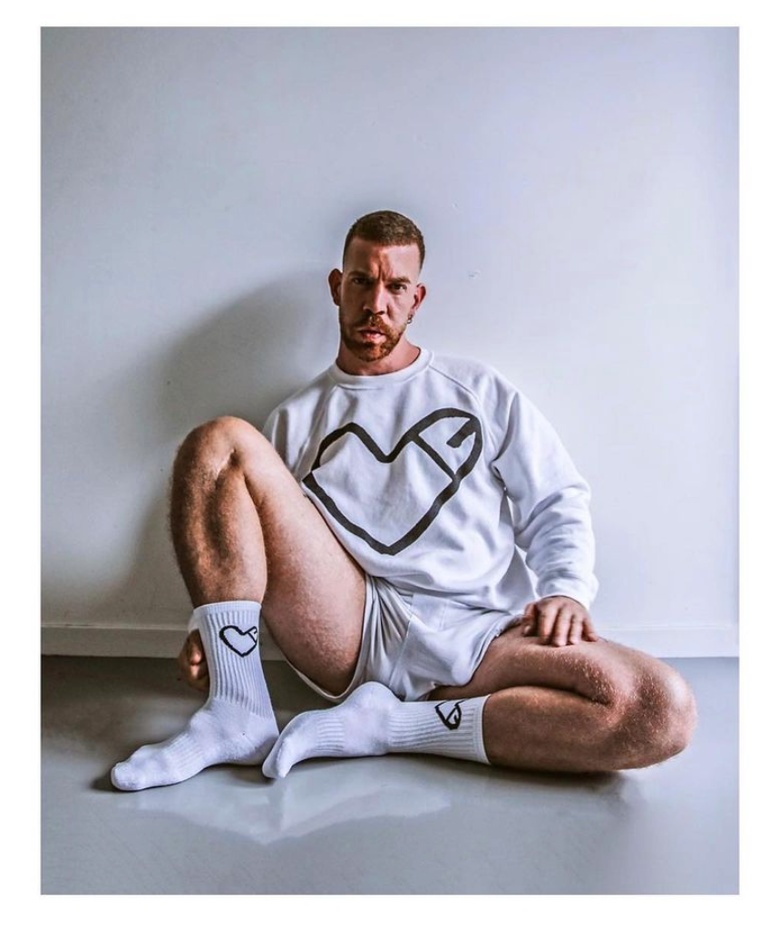 gay male with a large boner through his shorts wearing a grey sweatshirt and grey sweat socks with phallic illustrations by cockheart