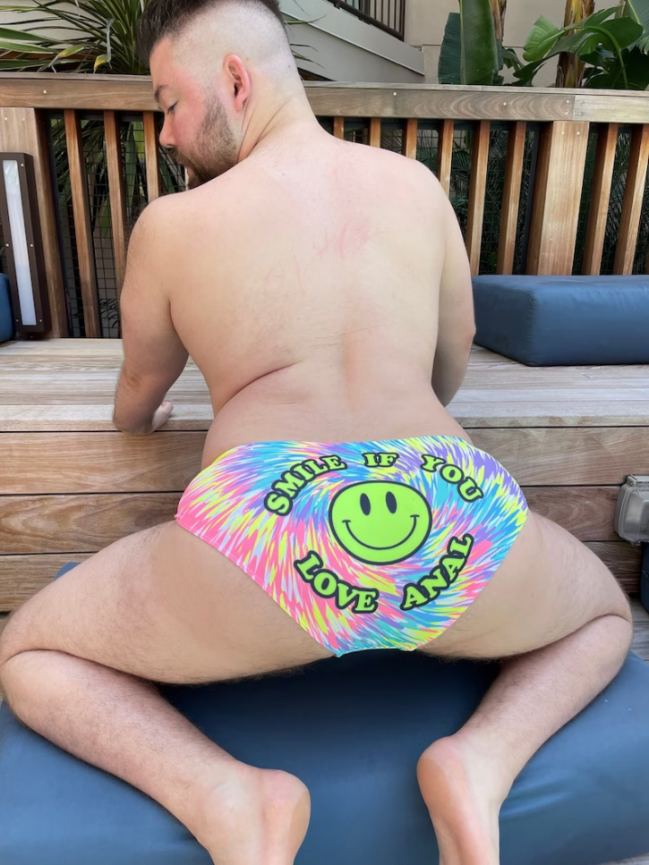 gay chubby bear kneeling on all fours in a tie dye speedo with a green smiley face that says smile if you love anal by Manny Valdez