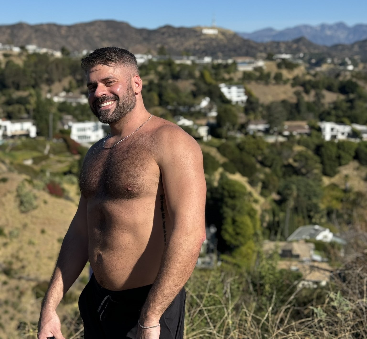 Max Romano standing shirtless outside smiling for a photo while wearing black sweat shorts