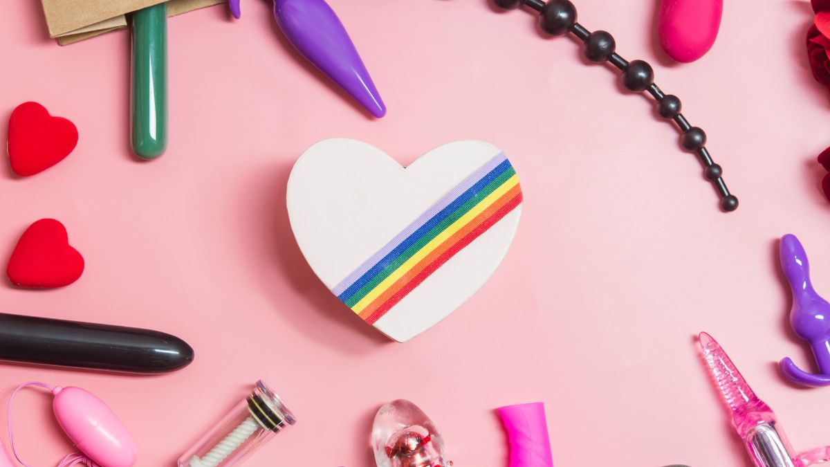 gay sex toys like anal beads dildos and butt plugs on a pink surface surrounded by a gay LGBT heart