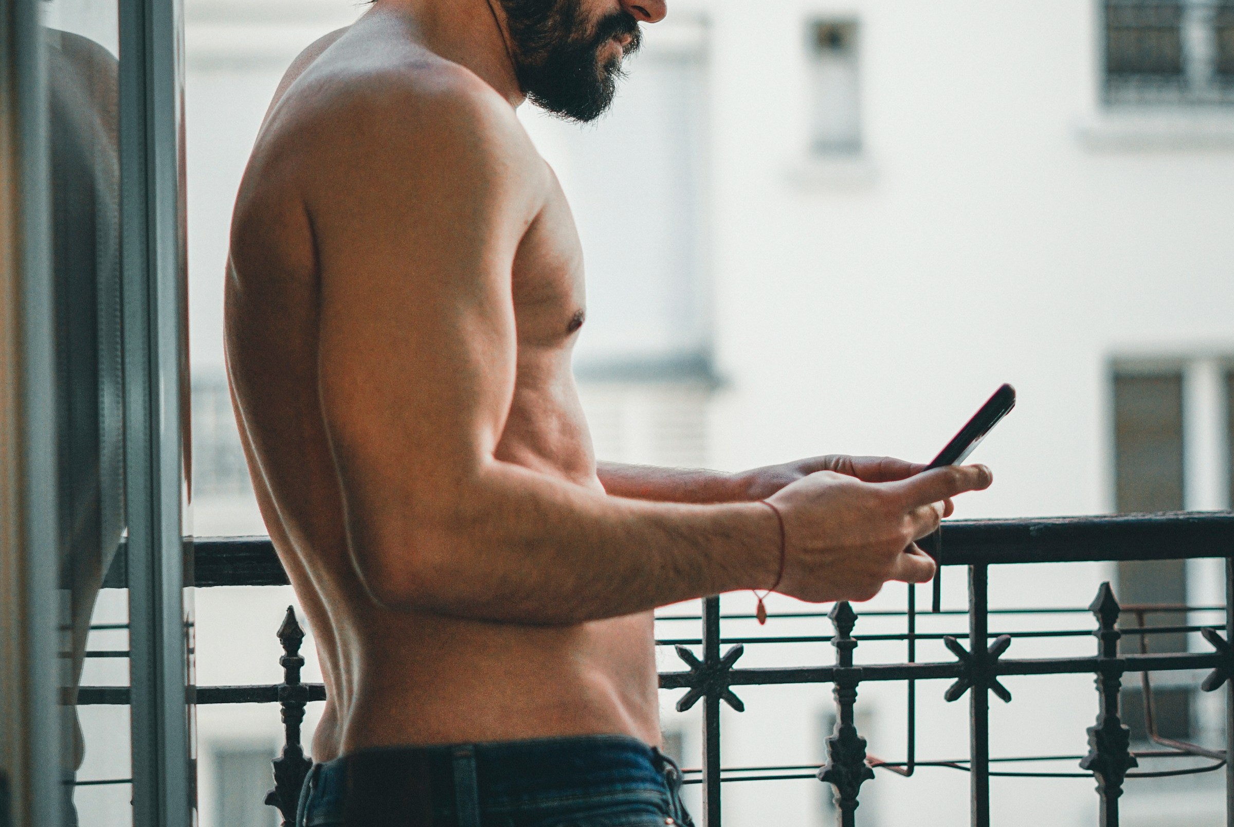 shirtless male on his phone looking at hot gay porn in jeans while standing outside on the balcony via thomas le-gourrierec unsplash