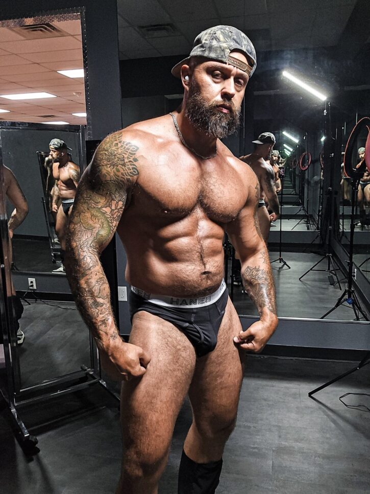 Gay porn star and OnlyFans creator Farm Dad flexing in black briefs and camo cap in a gym mirror, showing off his thick, tattooed muscle build