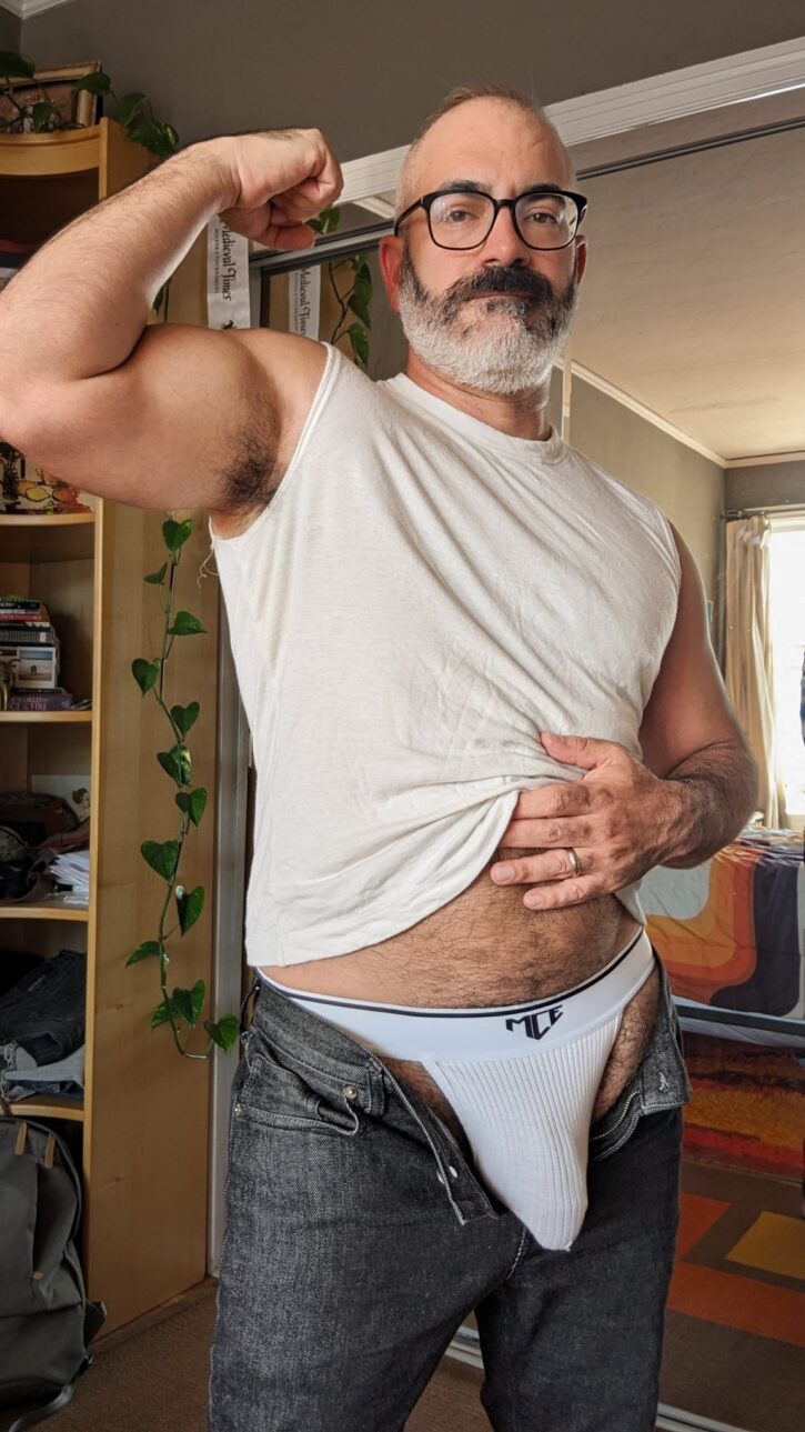 Fuzzz80 flexing in a white tank top and jockstrap, showing off his hairy chest, muscular arms, and daddy vibes. Gay porn creator and content model featured on JustForFans and OnlyFans.